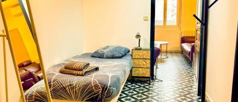 A narrow room with a bed, a small table, a chair, and a mirror.