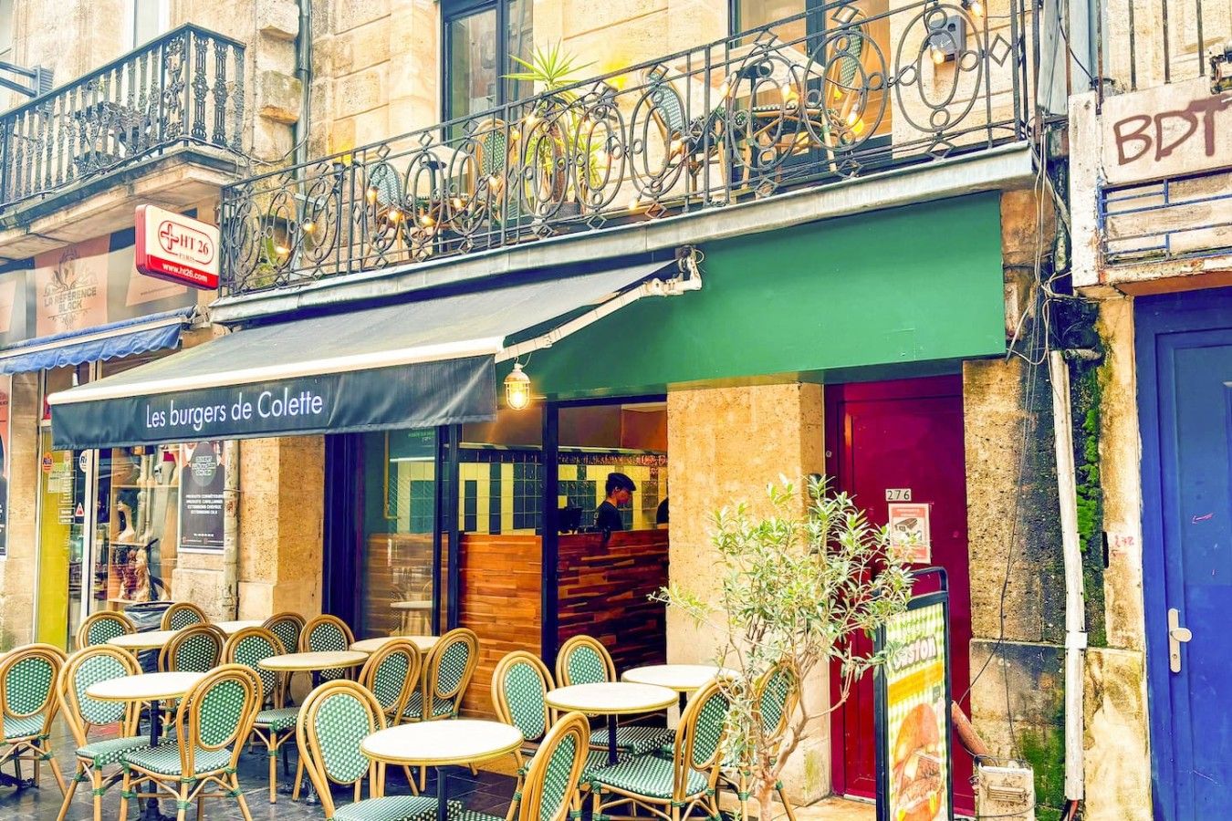 Un café en plein air avec des chaises et des tables en osier, un espace de repos couvert et un bâtiment surmonté d’une marquise verte.