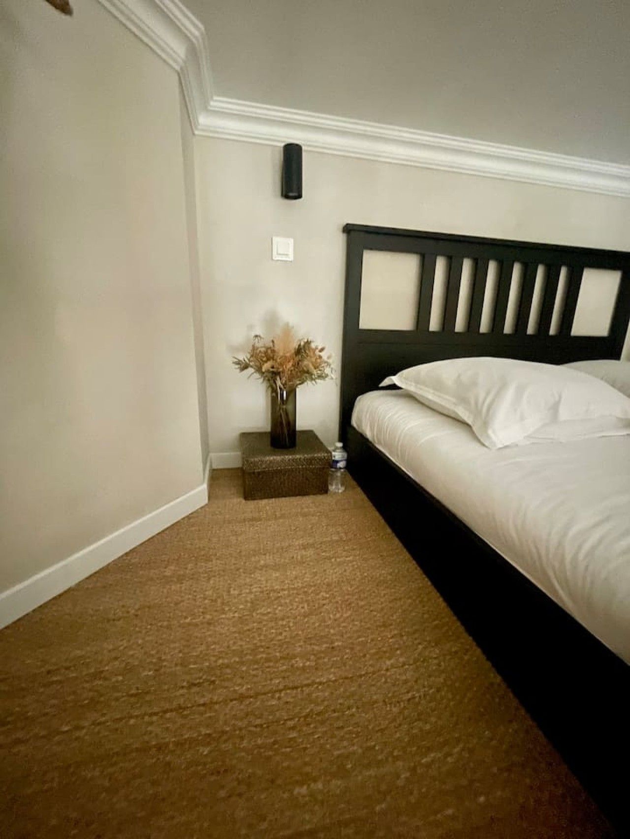 A bedroom with a bed, a nightstand, a vase with dried flowers, and a water bottle.