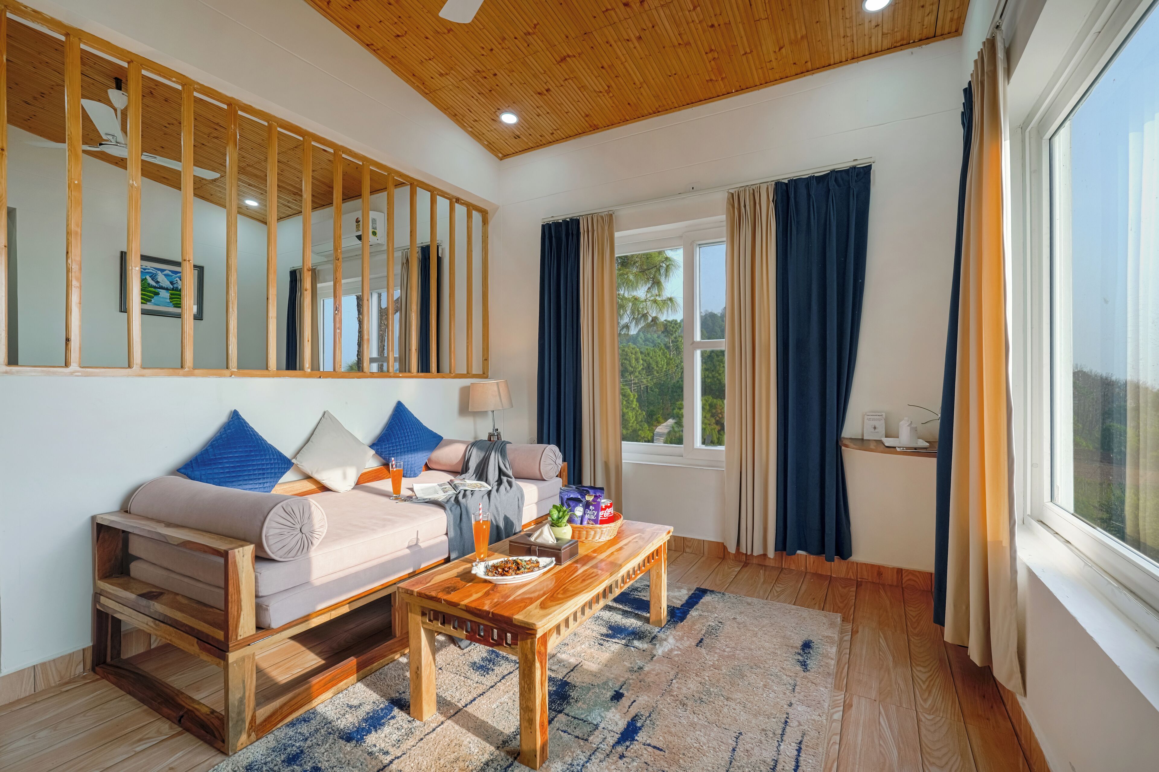 A cozy living room with a wooden ceiling, a sofa with cushions, a coffee table, and a large window with curtains.