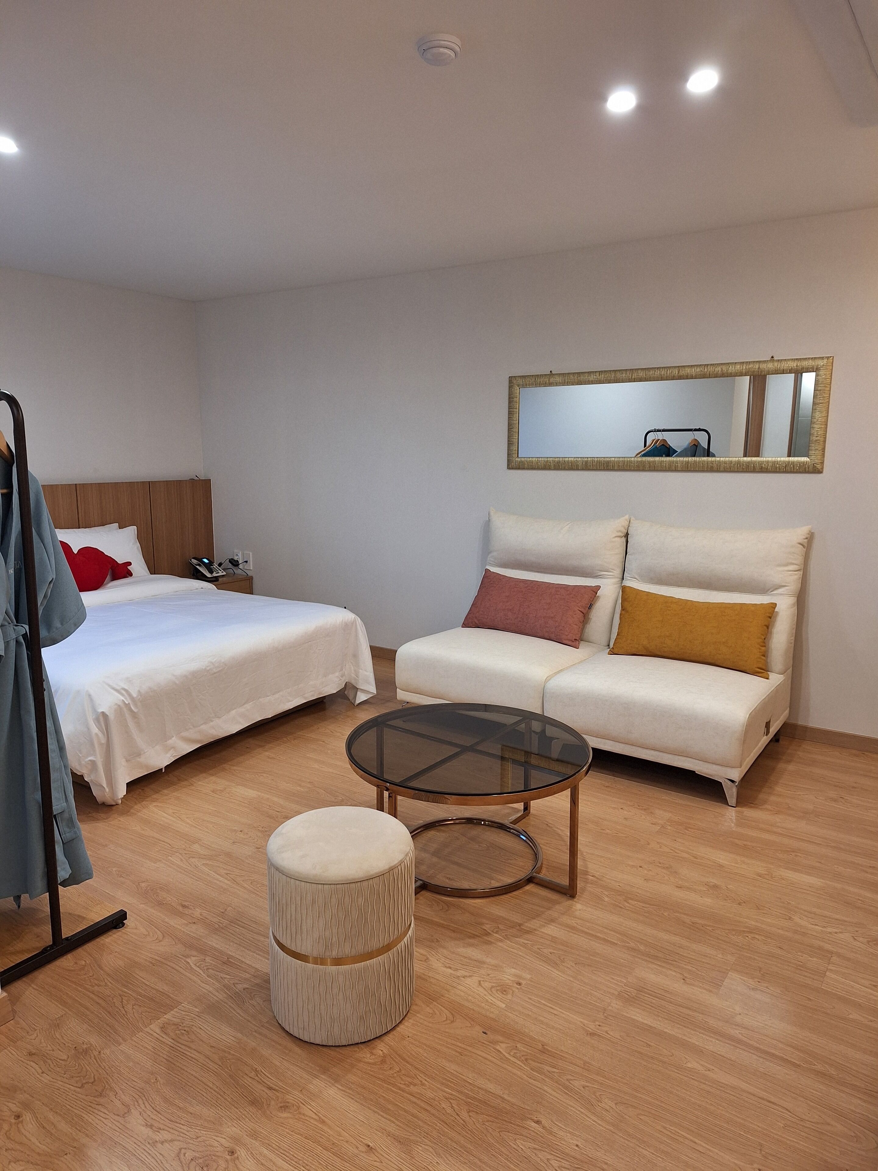 A hotel room with two beds, a sofa, a round glass coffee table, a small ottoman, a mirror, and a coat rack.