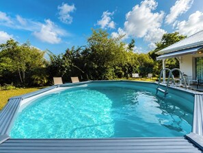 Una piscina blu cristallina con una scaletta metallica, circondata da vegetazione e un edificio bianco con un portico coperto.