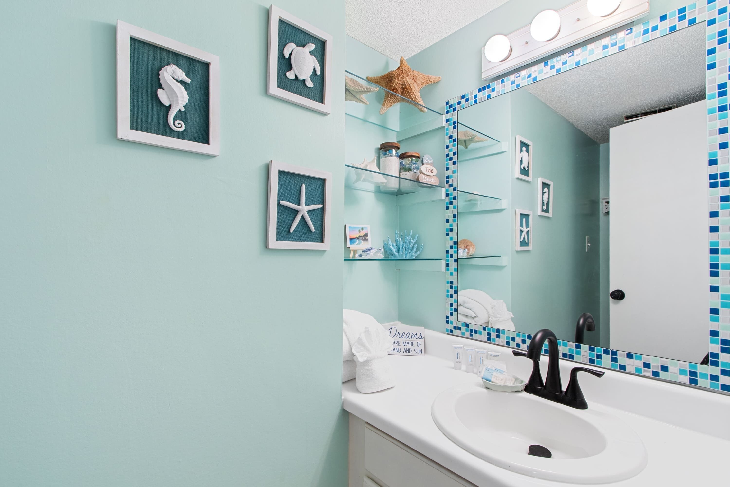 A bathroom with a teal wall, framed seahorse and starfish artwork, a glass shelf with toiletries, and a mirror with a blue-tiled border.