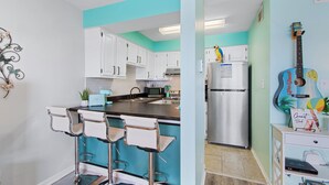 Een keuken met witte kasten, een blauw kookeiland met barstoelen, een roestvrijstalen koelkast en een gitaar aan de muur.