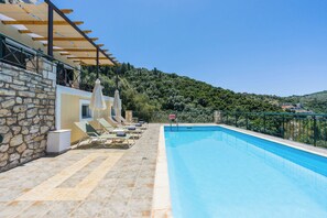 A swimming pool with lounge chairs, a stone wall, and a view of a green hillside. - Luxury seafront plot villa, seaviews, p-pool, walk to beach- Villa Alex Meganisi (Meganisi)