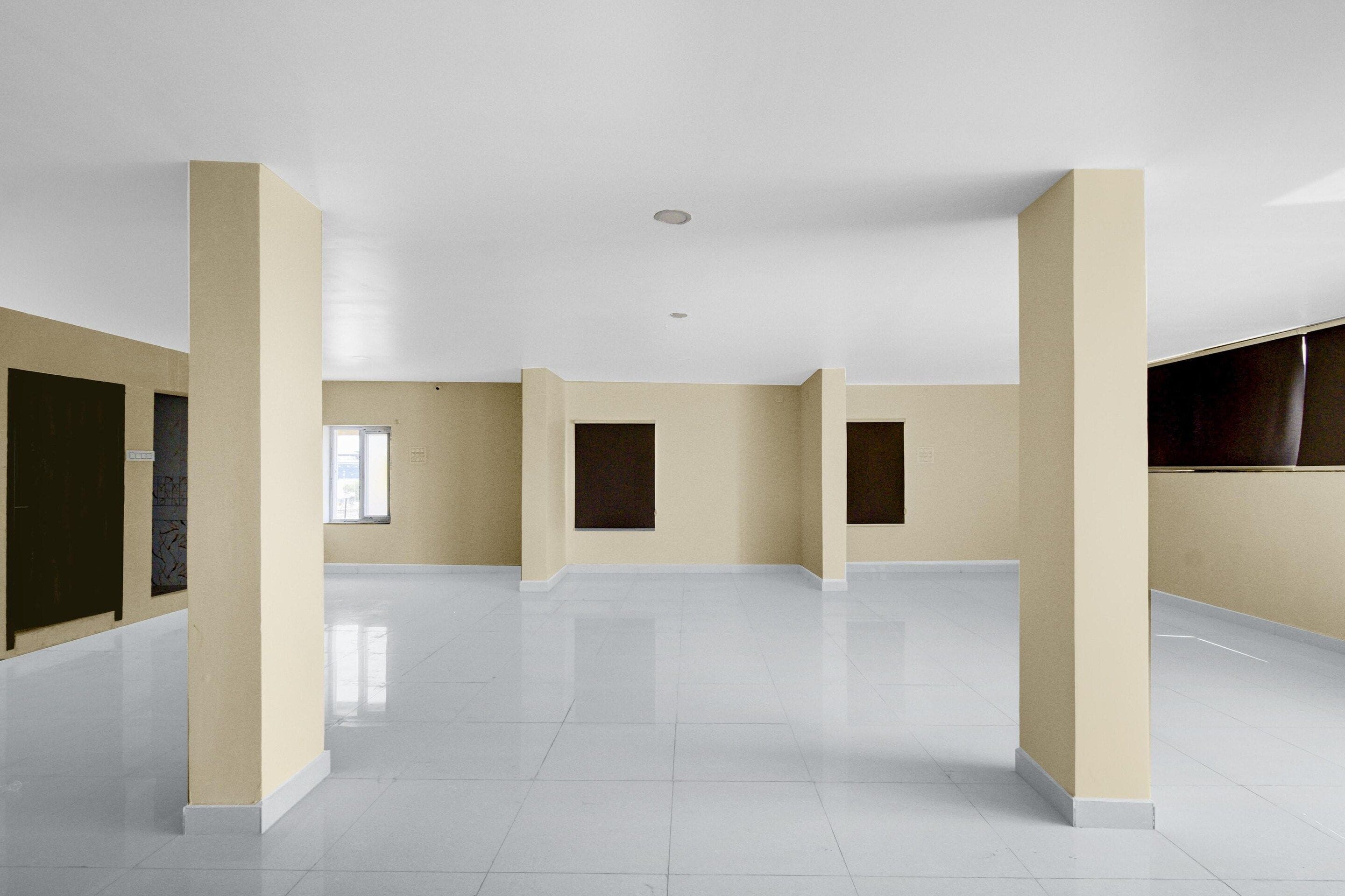 Ruime, lege kamer met witte betegelde vloeren en beige wanden, met grote ramen en een vlak plafond.