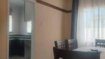 Een eethoek met een tafel en stoelen, een keuken op de achtergrond, en een raam met gordijnen.