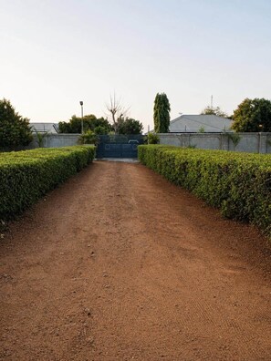 A dirt path leading to a gated entrance with a metal gate, flanked by neatly trimmed hedges.