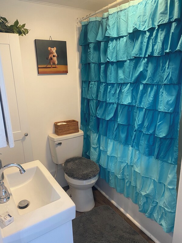Un baño con un lavamanos blanco, un inodoro con una cesta encima y una cortina de ducha azul con vuelo, además de un cuadro en la pared.