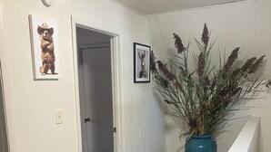 Een gang met houten vloer, witte muren en een blauwe vaas met planten. Twee ingelijste foto's hangen aan de muur.