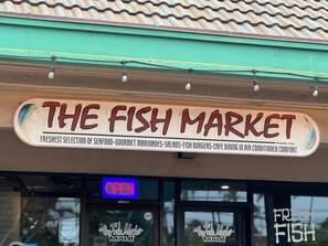 Una fachada con un letrero que dice "The Fish Market" que ofrece mariscos, adobos, ensaladas, hamburguesas de pescado y servicio de cafetería en un ambiente con aire acondicionado.