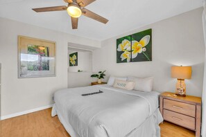 Une chambre à coucher comprenant un lit, une table de chevet, une lampe, un ventilateur de plafond et des œuvres d’art accrochées aux murs.