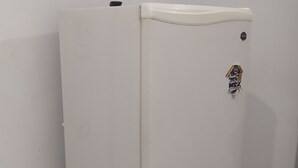 A white refrigerator with a sticker on the door, standing in a corner with a tiled floor and a plain wall.