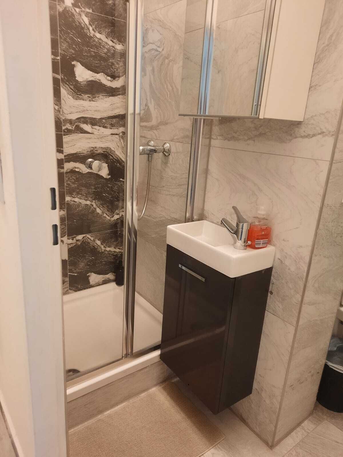 A bathroom with a glass shower enclosure, a white sink with a dark cabinet, and a marble backsplash.