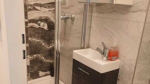 A bathroom with a glass shower enclosure, a white sink with a dark cabinet, and a marble backsplash. - Extended Stay 3 Budget Apartments (Vienna)