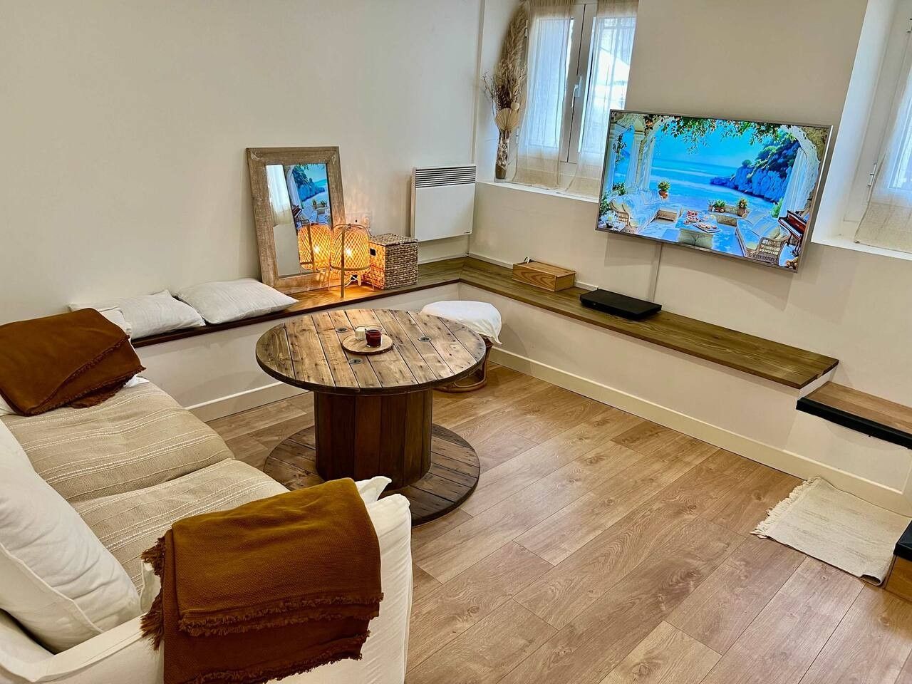 A living room with a round wooden coffee table, a sofa with cushions, a television mounted on the wall, and a window with a view.