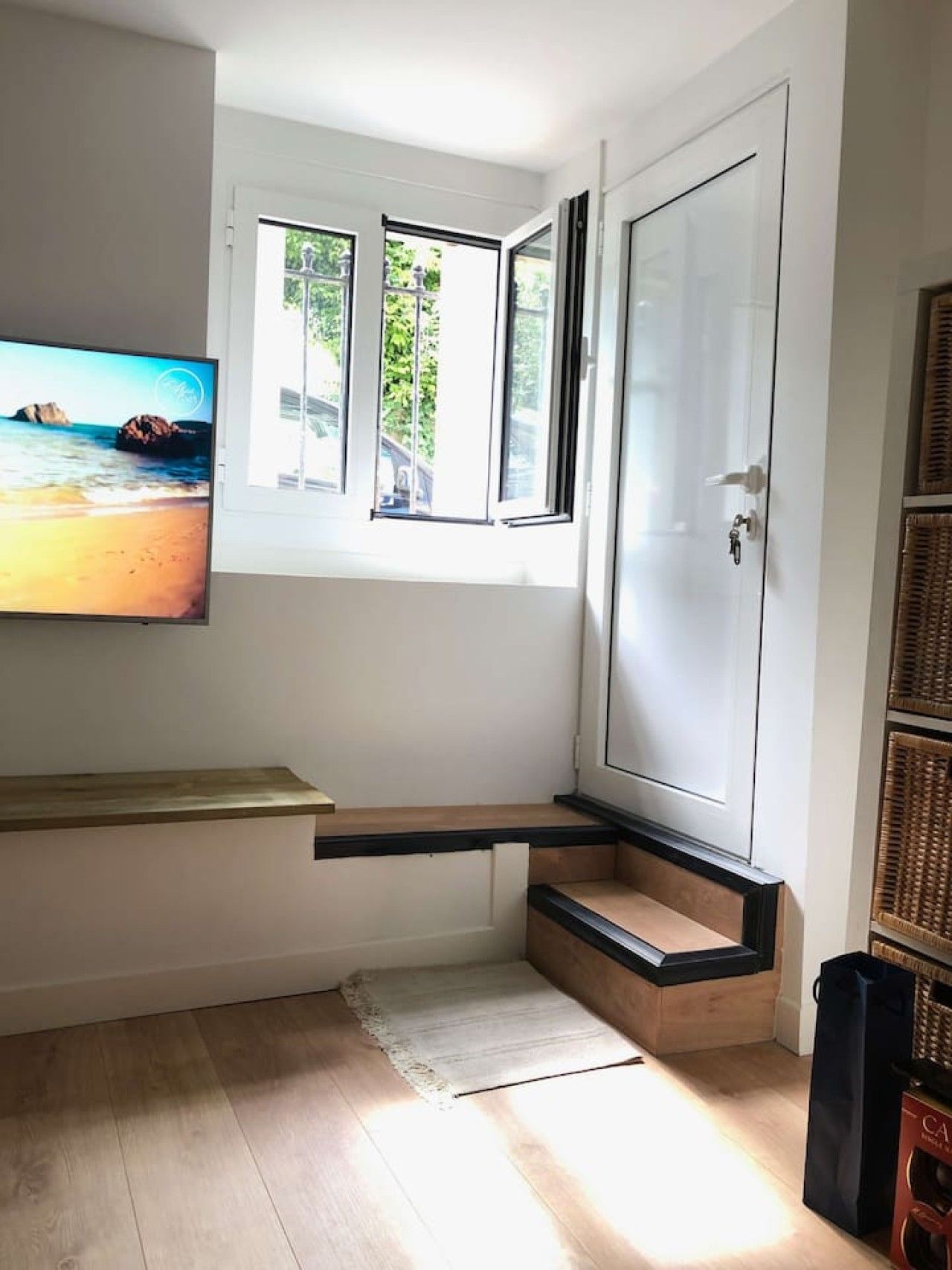 Ein Zimmer mit Fernseher an der Wand, einem Fenster mit Blick auf Grünflächen, einer Tür und einer kleinen Holzbank.