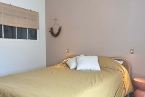 Ein Einzelbett mit beiger Bettdecke und weißen Kissen, ein Holz-Kopfteil, eine Wandleuchte und ein Fenster mit Jalousien.