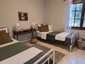 A room with two beds, a bedside table with lamps, a window with a view of trees, and a painting on the wall.