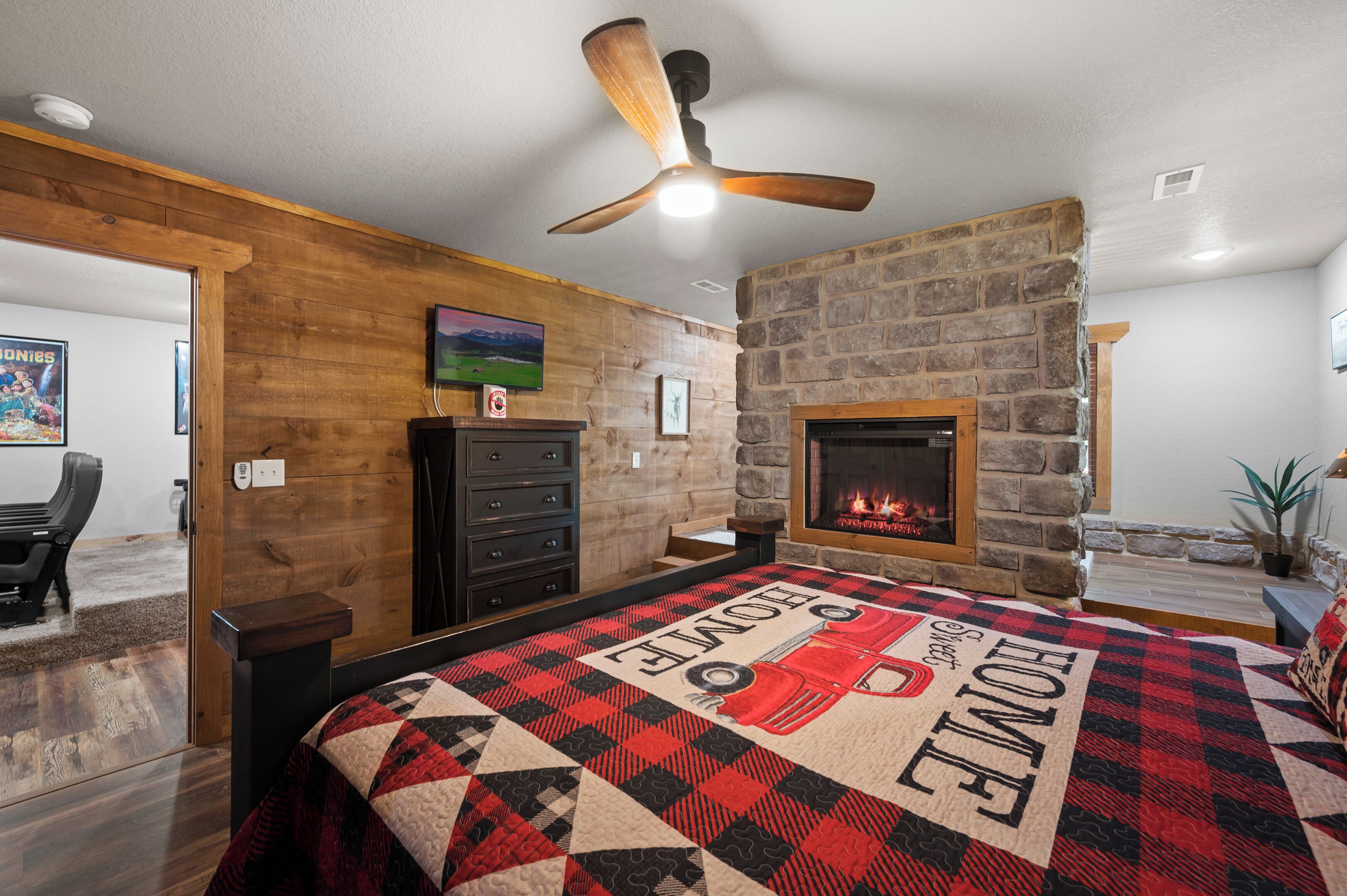 A bedroom with a fireplace, a bed with a quilt, a ceiling fan, and a stone wall.