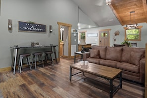 A modern interior with a kitchen area, bar stools, a brown sofa, and a wooden coffee table. - The Ultimate Staycation: Indoor Pool, Hot Tub & Game Room Cabin (Sevierville)