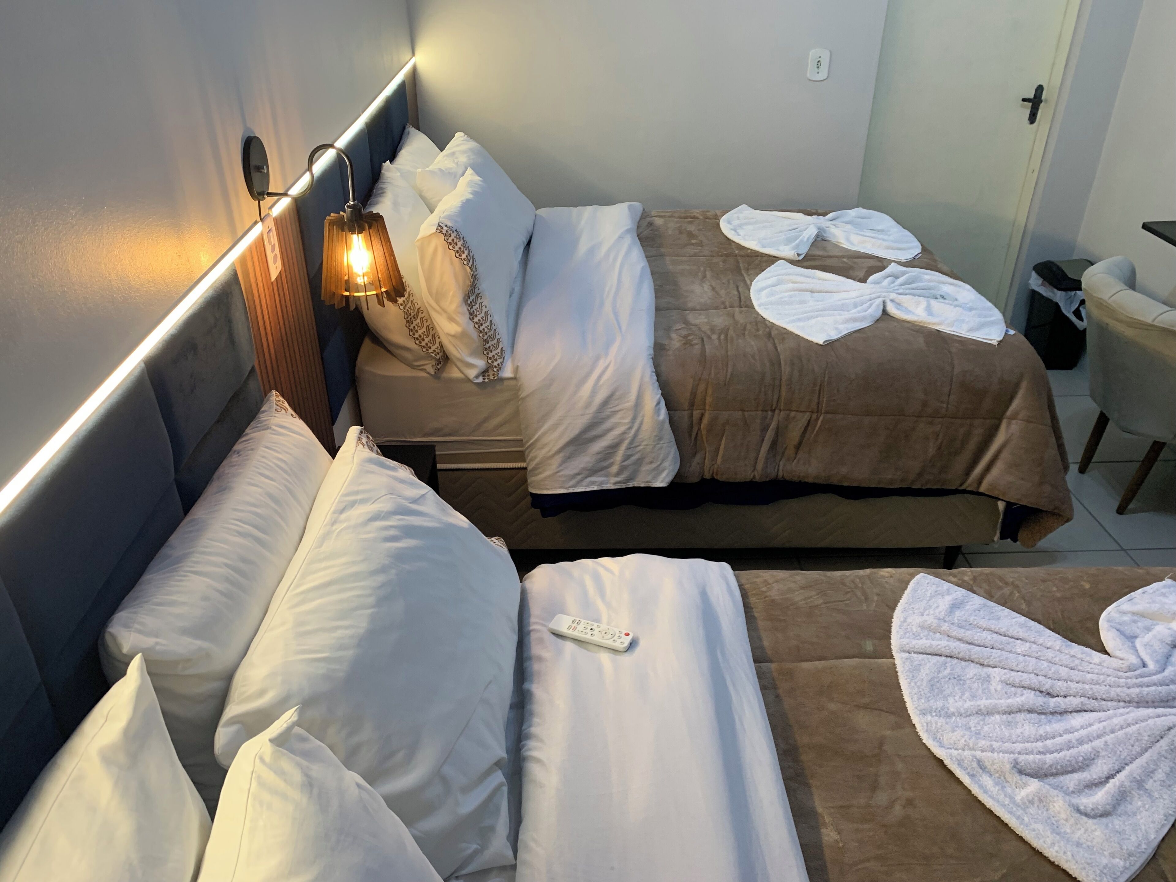 Quarto de hotel com duas camas, uma mesa de cabeceira com abajur, uma cadeira e uma porta.