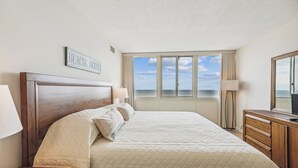 Habitación de hotel con una cama grande, cabecera de madera, mesita de noche, escritorio, lámpara, espejo y ventana con vista al océano.