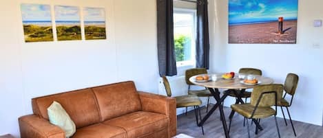 A furnished room with a brown leather sofa, a wooden coffee table, a dining table with chairs, and artwork on the walls.