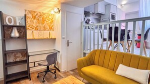 Ett kompakt bostadsutrymme med ett skrivbord, en stol, en soffa och ett köksområde som syns genom en glasdörr.