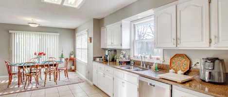 Una cocina con armarios blancos, una mesa de comedor con sillas, una cafetera y un lavavajillas.