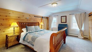 Un dormitorio con cabecera de madera, cama con edredón estampado con flores, mesita de noche con lámpara y ventana con cortinas.