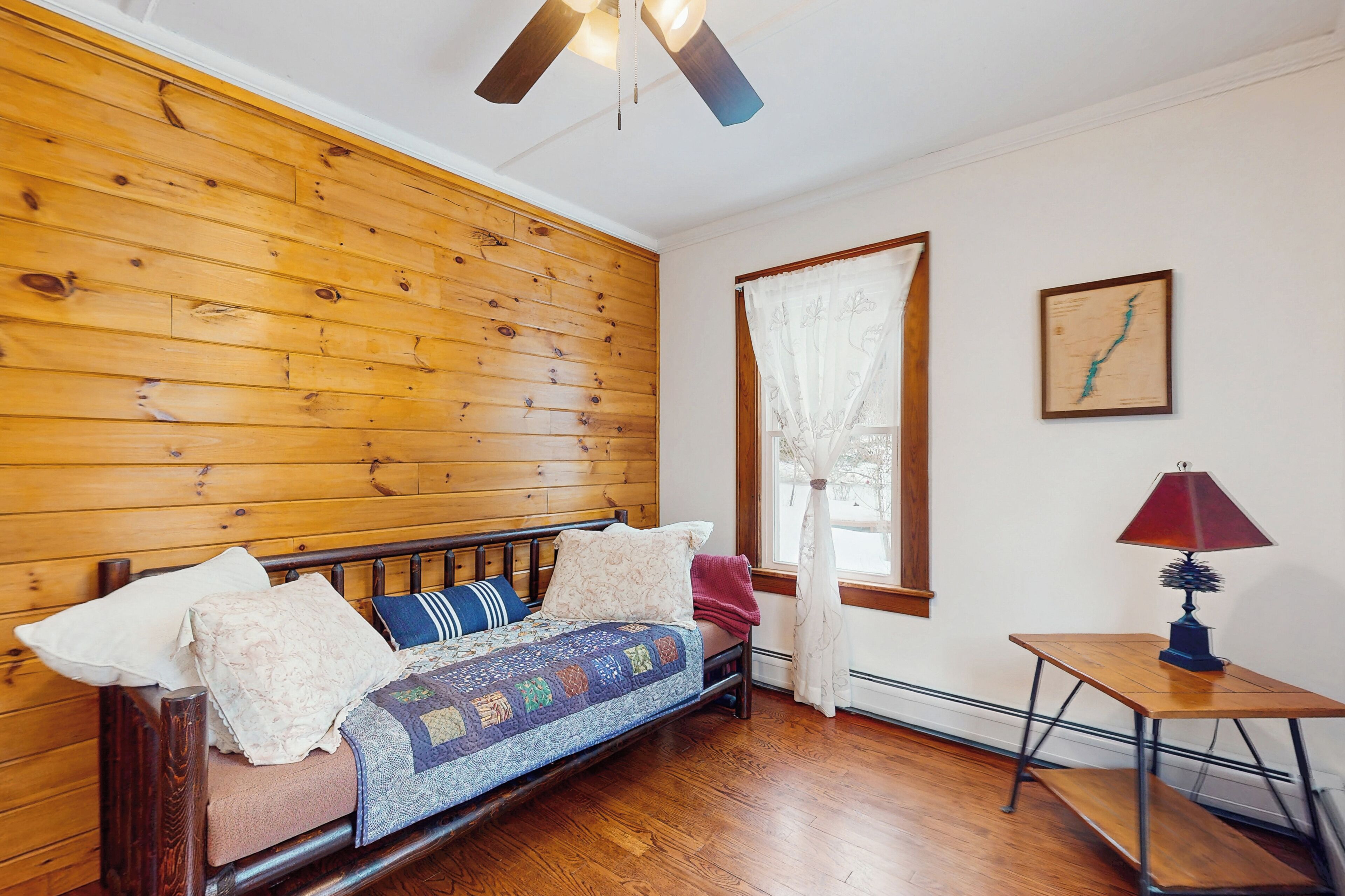 A cozy room with a wooden wall, a bed with a quilt, a small table with a lamp, and a window with curtains.