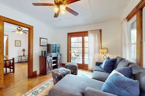 A living room with a gray sofa, ottomans, a ceiling fan, a wooden floor, a TV, and a window with curtains. - 2 Mins to Lake Park, Beach & Town! Cozy Cottage (Bolton Landing)