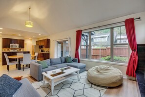 A modern living room with a grey sofa, a white coffee table, and a bean bag. There is a kitchen area with a microwave and cabinets, and a dining table with chairs.