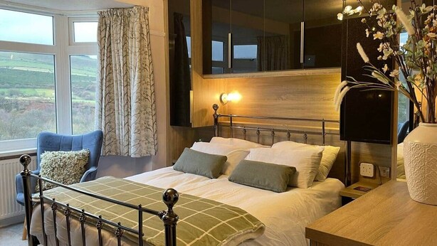 Un dormitorio con cama, mesita de noche, lámpara, silla y vistas al exterior.