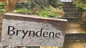 Un letrero de madera con el nombre 'Bryndene' frente a un edificio de ladrillo rojo con ventanal saliente y puerta blanca.