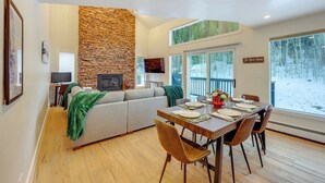 Una sala de estar acogedora con una chimenea de piedra, un comedor con mesa y sillas de madera, y un amplio ventanal con vistas a un paisaje nevado.