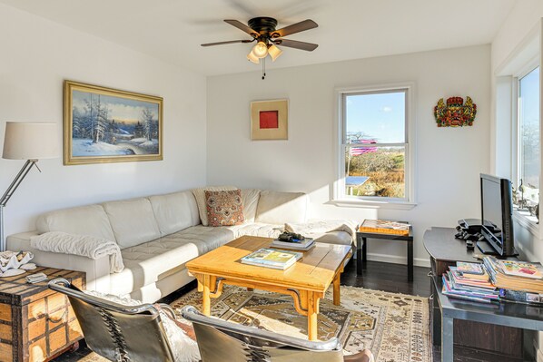 A living room with a sofa, coffee table, and a television. There is a ceiling fan, a window with a view, and a painting on the wall. - 1 Block to Bellport Bay! Scenic Hamptons Hideaway (Shirley)
