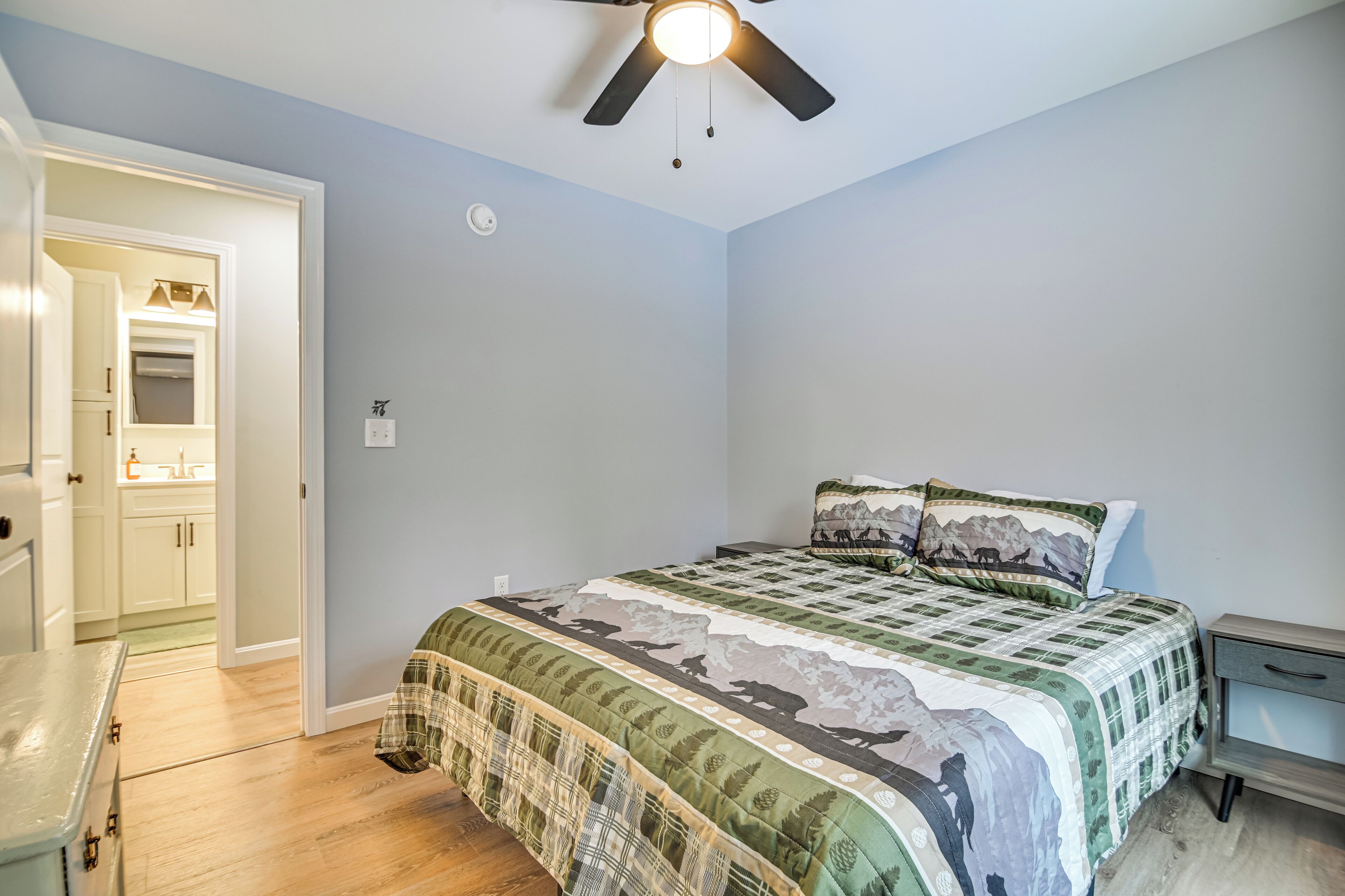A bedroom with a bed, a nightstand, a ceiling fan, and a door leading to a bathroom.
