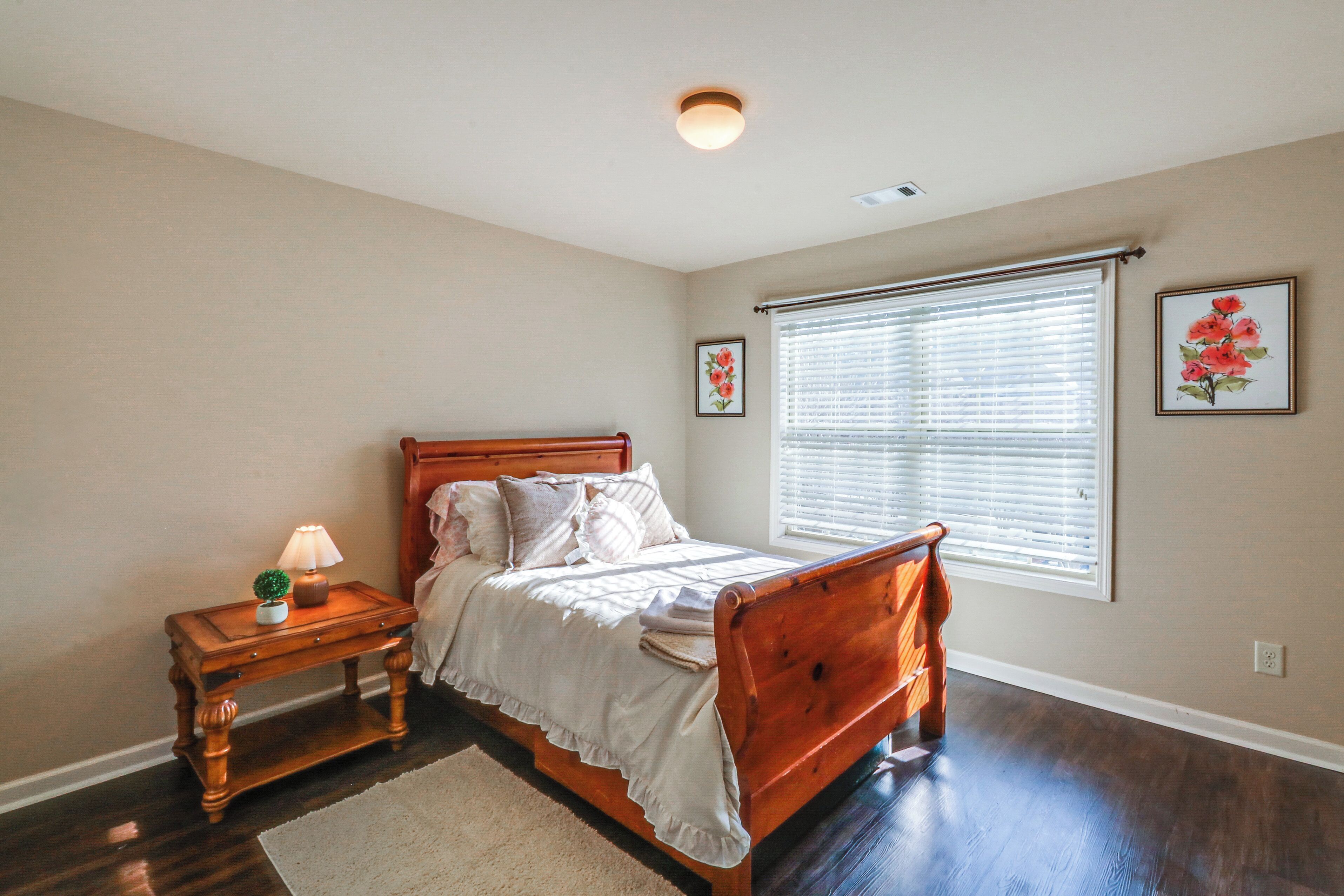 A bedroom with a wooden bed, a nightstand with a lamp, a window with blinds, and two framed pictures on the wall.