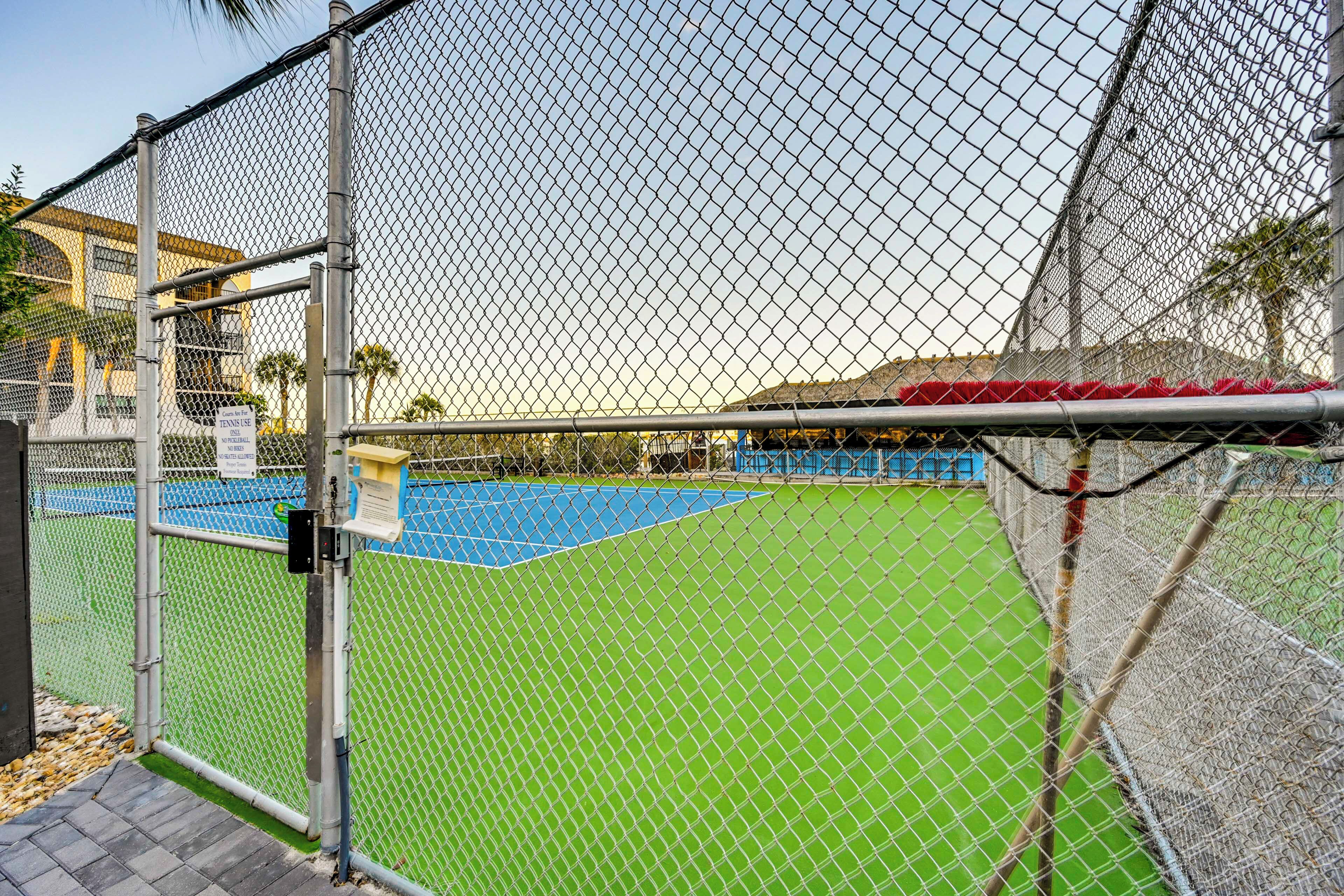 A chain-link fence surrounding a tennis court with a green surface and a blue area, possibly a pool or water feature.