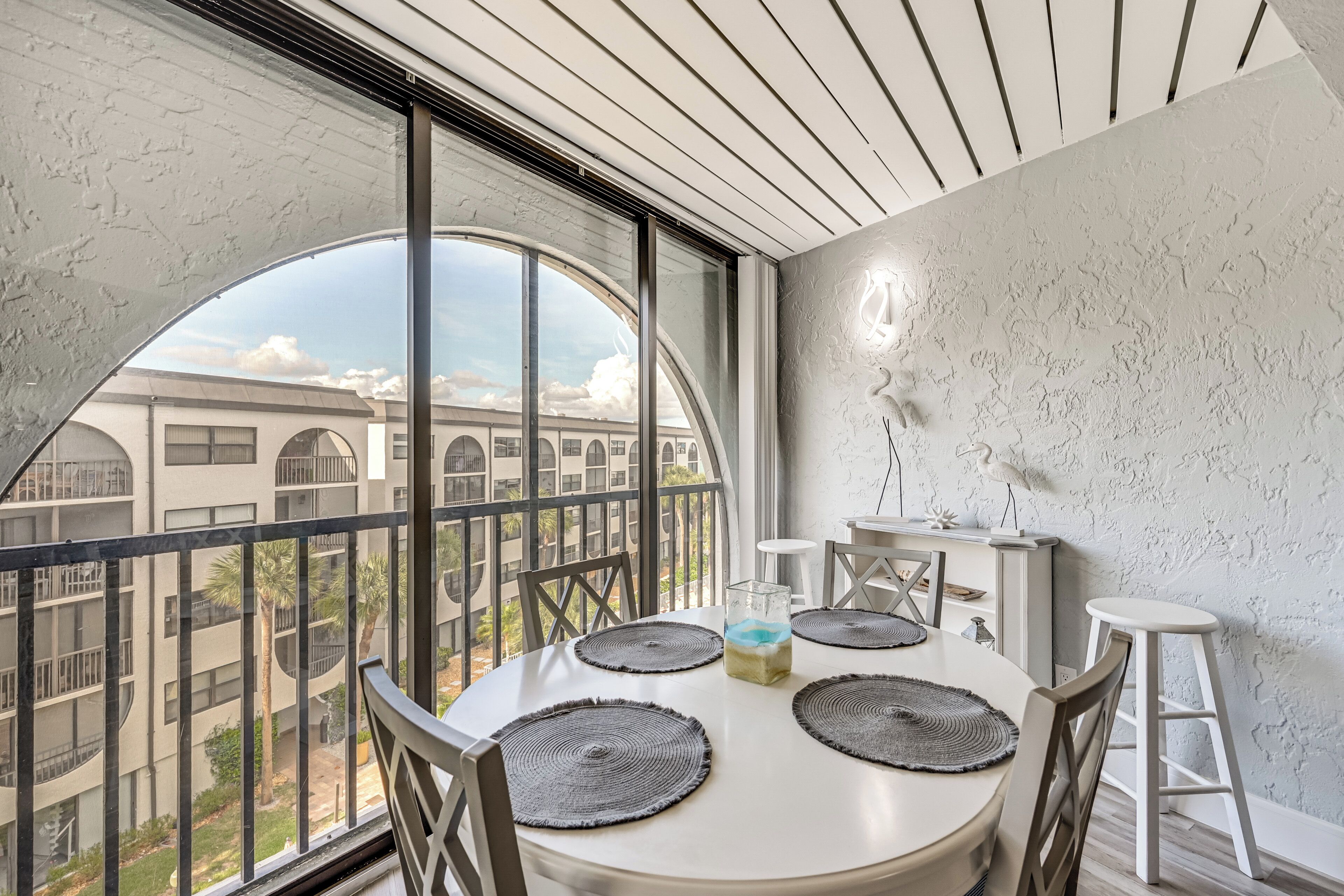 A dining area with a round table, four chairs, a glass of water, and a view of a building through large windows.