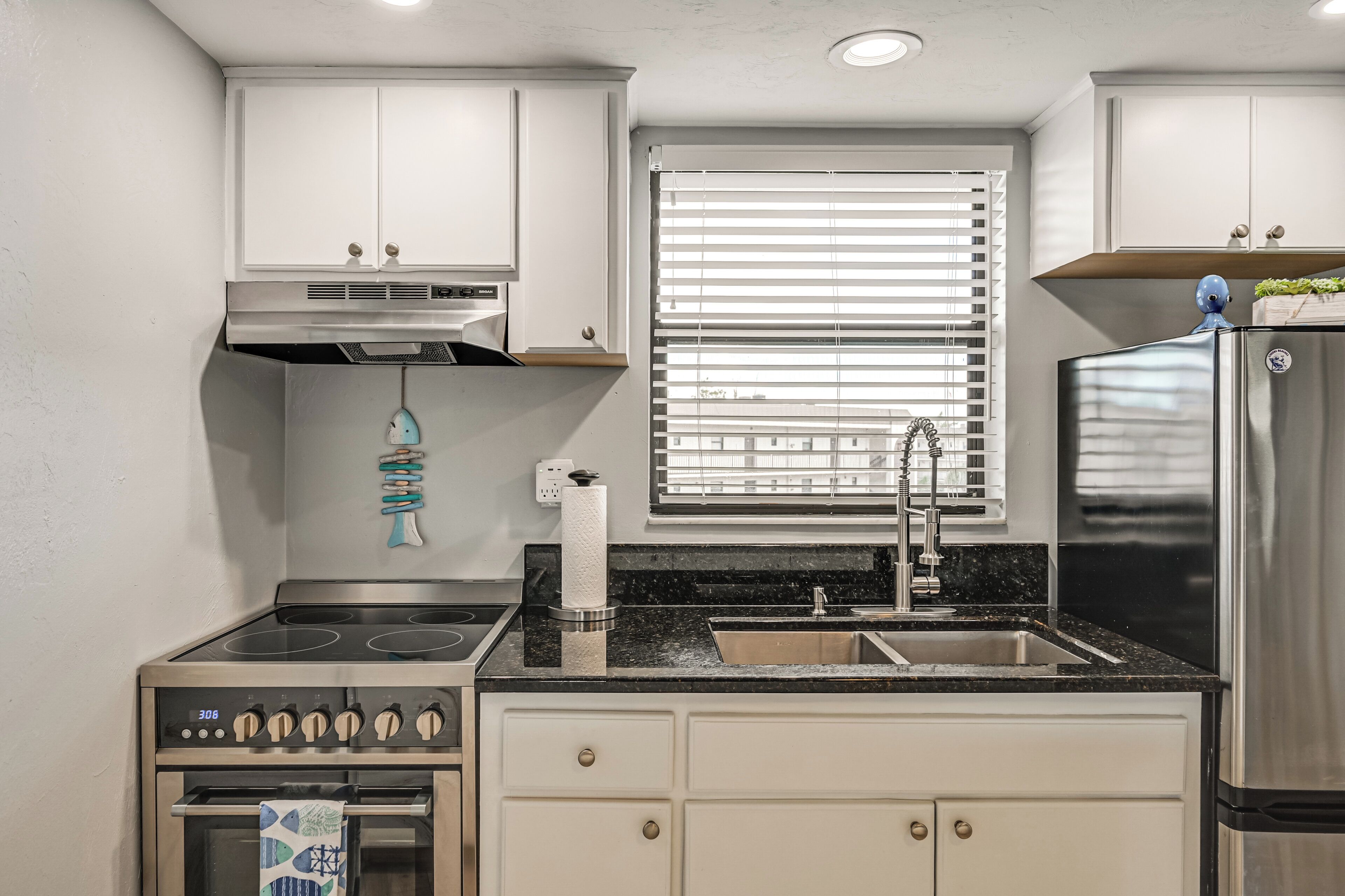 Una cocina moderna con armarios blancos, un refrigerador de acero inoxidable, un horno, un lavamanos y una ventana con persianas.