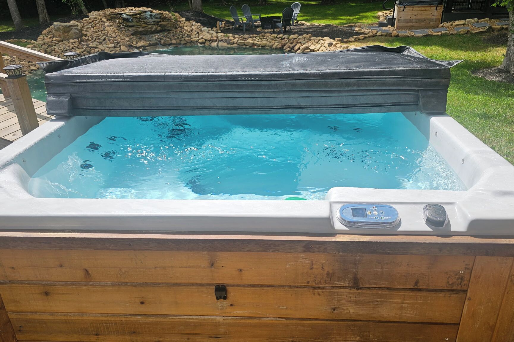 A hot tub with a control panel, surrounded by a wooden deck and a stone border.