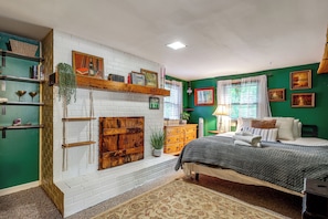 Un dormitorio con chimenea de ladrillo blanco, paredes verdes, una cama con manta gris y un aparador de madera.