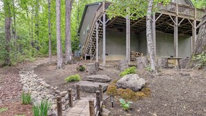A rustic cabin with a wooden deck, surrounded by a forested area with a stone pathway leading to the entrance. - Modern Mountain Zen 3-bed, 3-bath Home With Creeks (Sapphire)