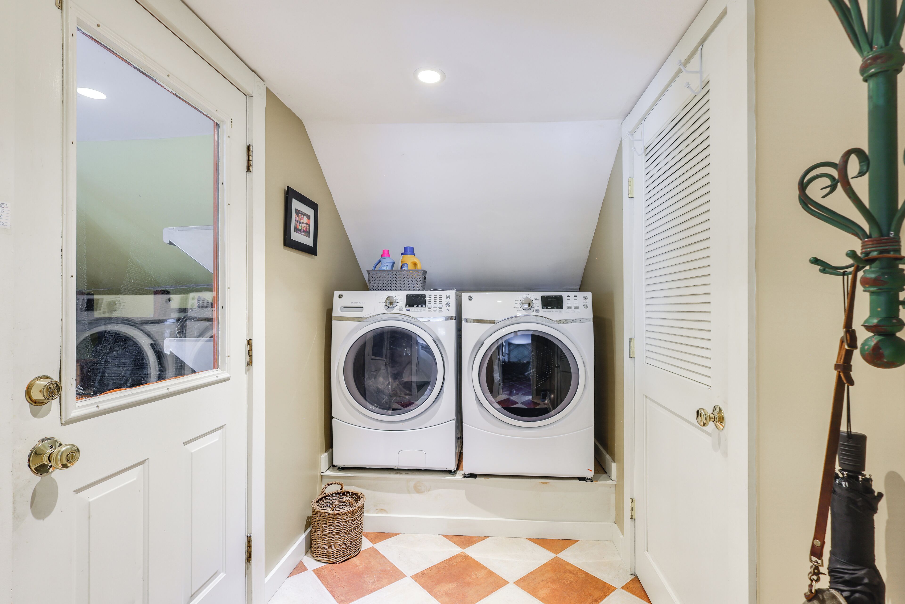 A laundry room with two stacked front-loading washing machines, a wicker basket, and a door with a glass panel.