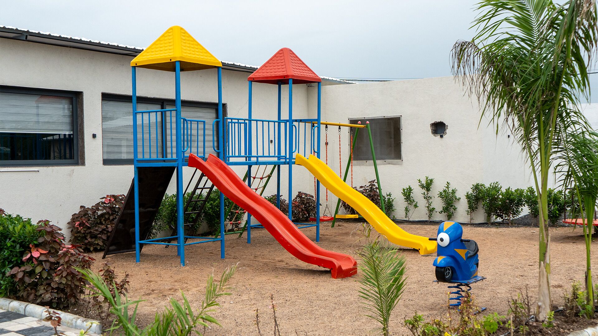 A playground with slides and a climbing structure, surrounded by a sandy area and plants.