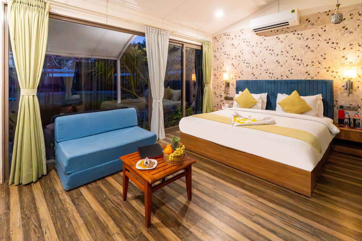 Ein Schlafzimmer mit einem großen Bett, einem blauen Sofa, einem Holztisch mit Obstschale und Blick durch eine Schiebeglastür.