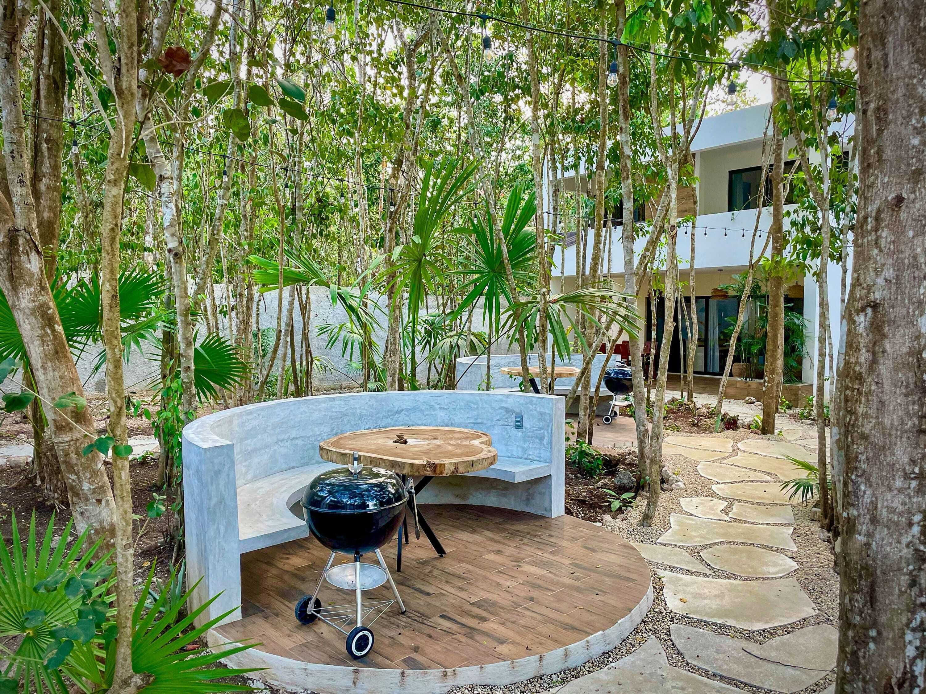 A circular wooden deck with a barbecue grill, surrounded by trees and a stone pathway.
